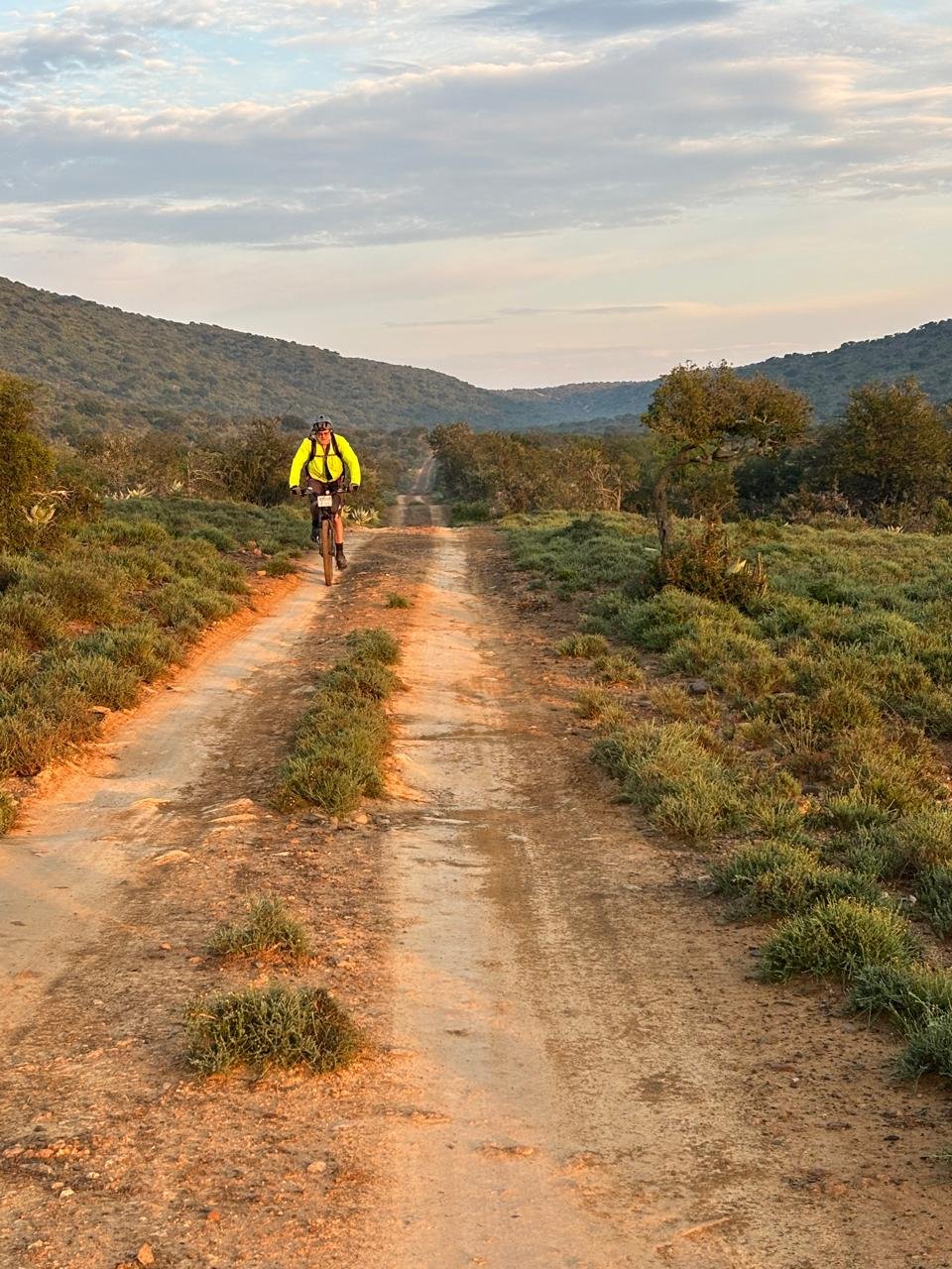 A rider on a red gravel track through the bush at dawn, with hills behind